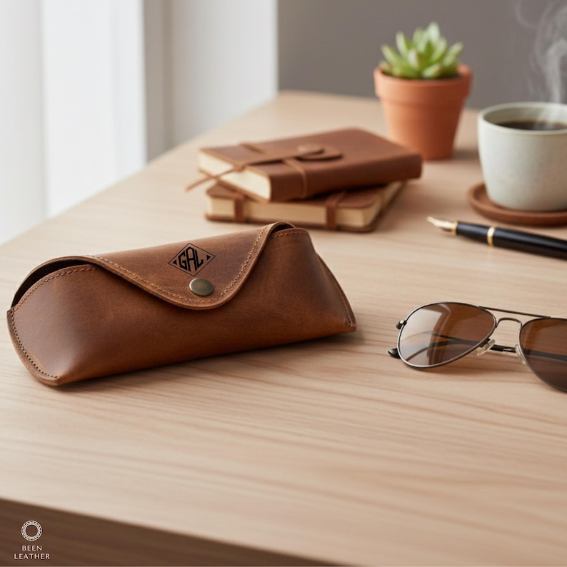 May include: A brown leather glasses case with the initials "GAL" embossed on the flap, alongside aviator sunglasses. A small succulent in a terracotta pot, a cup of coffee, and two leather-bound notebooks are on the wooden surface.