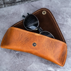 May include: A tan leather sunglasses case with a snap closure, containing black sunglasses. The case is open, showing a dark brown interior. The case has a textured surface.
