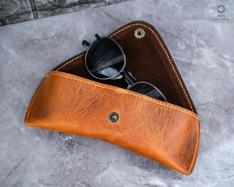 May include: A tan leather sunglasses case with a snap closure, containing black sunglasses. The case is open, showing a dark brown interior. The case has a textured surface.