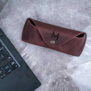 May include: A burgundy leather glasses case with a snap closure. The case is personalized with the initials "M" and the name "MICHAEL" in black lettering. The case is on a grey marble surface next to a laptop.