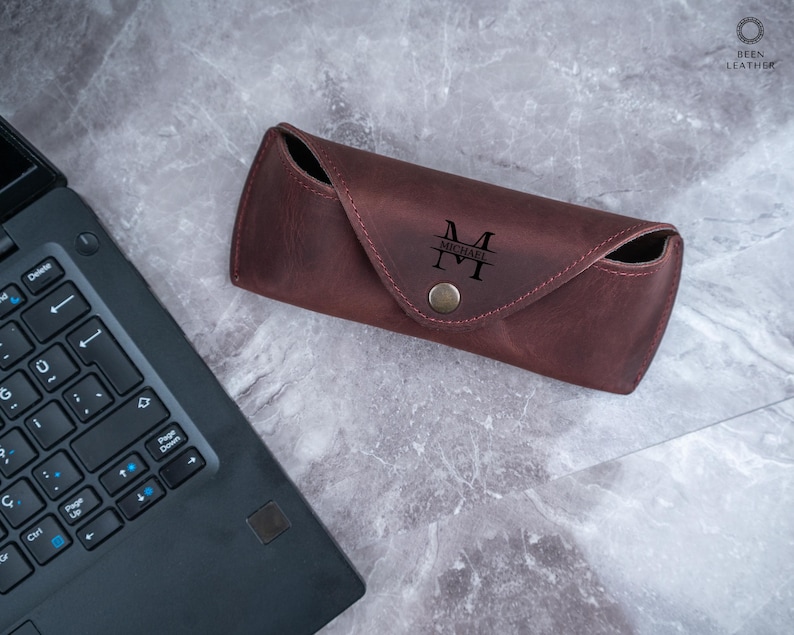 May include: A burgundy leather glasses case with a snap closure. The case is personalized with the initials "M" and the name "MICHAEL" in black lettering. The case is on a grey marble surface next to a laptop.