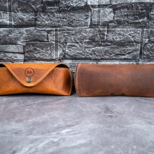 May include: Two brown leather eyeglass cases. One case is a trapezoid shape with a snap closure and the initials "BA" in a circle. The other case is a cylindrical shape. Both cases are made of textured brown leather.
