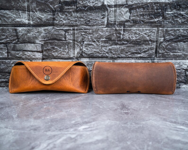 May include: Two brown leather eyeglass cases. One case is a trapezoid shape with a snap closure and the initials "BA" in a circle. The other case is a cylindrical shape. Both cases are made of textured brown leather.