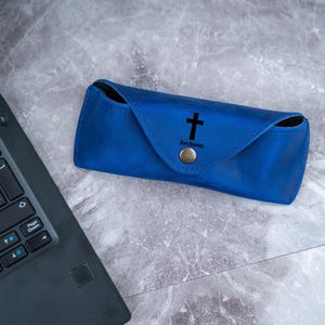 May include: A cobalt blue leather glasses case featuring a black cross and the name "Jackson". The case has a snap closure. It is on a marble surface next to a laptop.