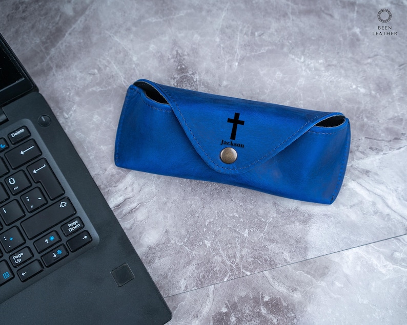 May include: A cobalt blue leather glasses case featuring a black cross and the name "Jackson". The case has a snap closure. It is on a marble surface next to a laptop.