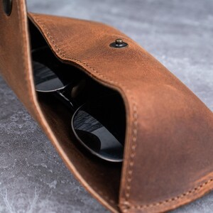 May include: A brown leather sunglasses case, open to show a pair of sunglasses. The case has a snap closure and visible stitching. The sunglasses have dark lenses and reflect the surrounding environment.