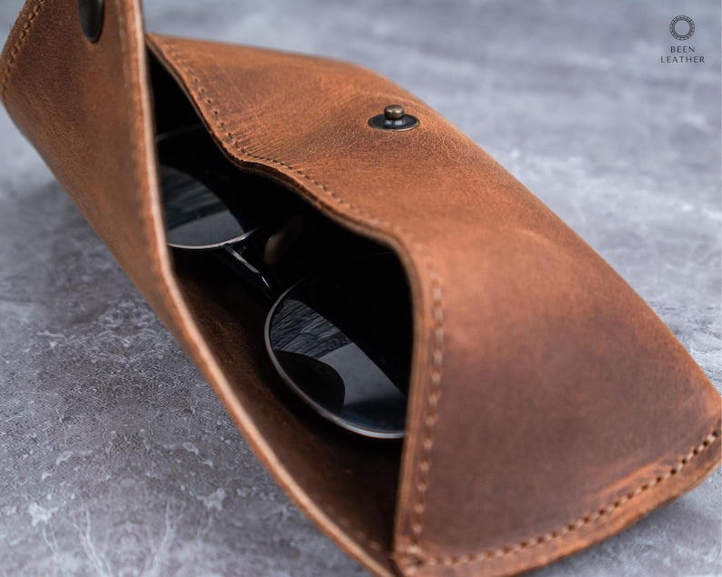 May include: A brown leather sunglasses case, open to show a pair of sunglasses. The case has a snap closure and visible stitching. The sunglasses have dark lenses and reflect the surrounding environment.