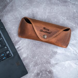 May include: A brown leather glasses case with the text "Merry Christmas!" embossed on the front. The case has a snap closure and is on a grey marble surface. A laptop is partially visible in the image.