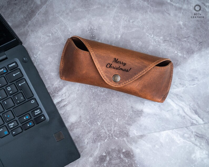 May include: A brown leather glasses case with the text "Merry Christmas!" embossed on the front. The case has a snap closure and is on a grey marble surface. A laptop is partially visible in the image.