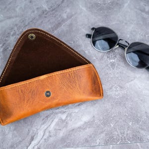 May include: A tan leather sunglasses case with a snap closure, open to show a dark brown interior. Round sunglasses with black frames and lenses are next to the case. The case has a distressed finish and visible stitching.