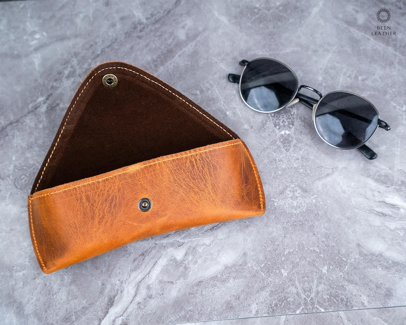May include: A tan leather sunglasses case with a snap closure, open to show a dark brown interior. Round sunglasses with black frames and lenses are next to the case. The case has a distressed finish and visible stitching.