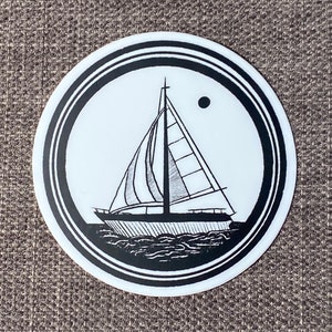 May include: Black and white sticker of a sailboat with a single mast and a full sail. The sailboat is in a circle with a black border and a white background. The sticker is on a brown fabric background.