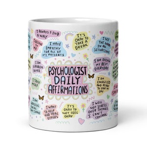 Op de afbeelding: Een witte keramische mok met kleurrijke tekst en illustraties. De tekst luidt "Psychologist Daily Affirmations" en bevat affirmaties zoals "Ik vind altijd een manier", "Ik heb empathie voor al mijn patiënten", "Het is oké om een ​​pauze te nemen", "Ik zie het goede in alle situaties", "Ik doe mijn best elke dag", "Ik ben energiek en klaar om mijn dag te beginnen", "Ik zal me niet druk maken over dingen die ik niet kan controleren".