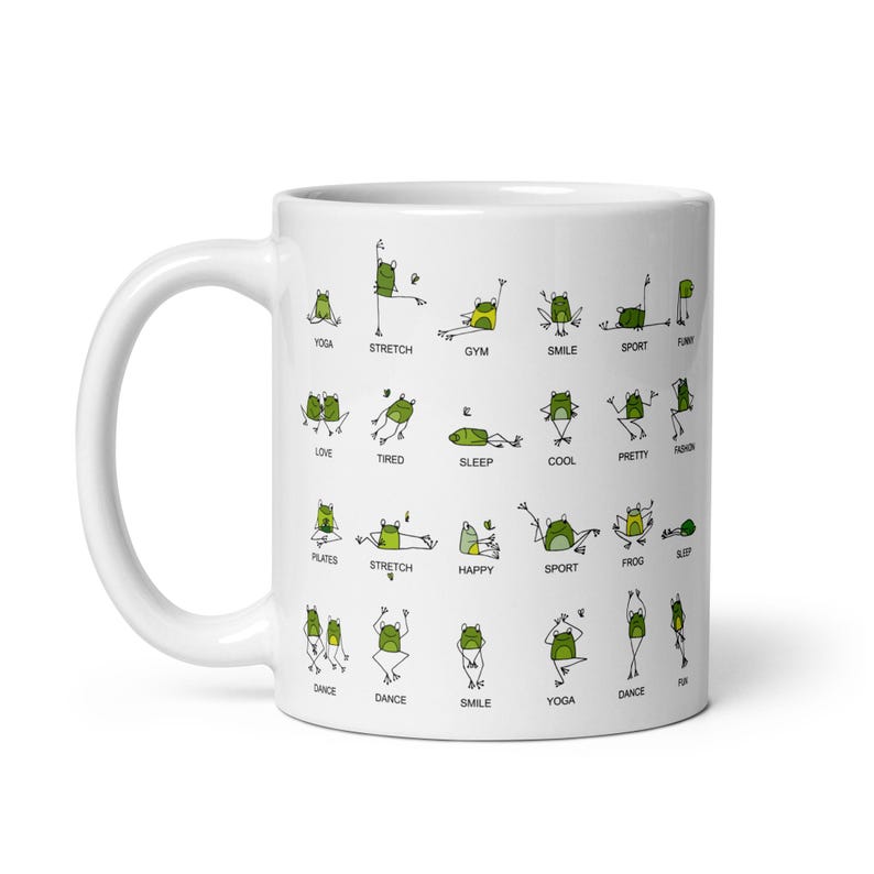 May include: White ceramic mug featuring a whimsical design of green frogs in various yoga poses. The mug has the words "YOGA", "STRETCH", "GYM", "SMILE", "SPORT", "LOVE", "TIRED", "SLEEP", "COOL", "PRETTY", "PLATES", "HAPPY", "FROG", and "DANCE".