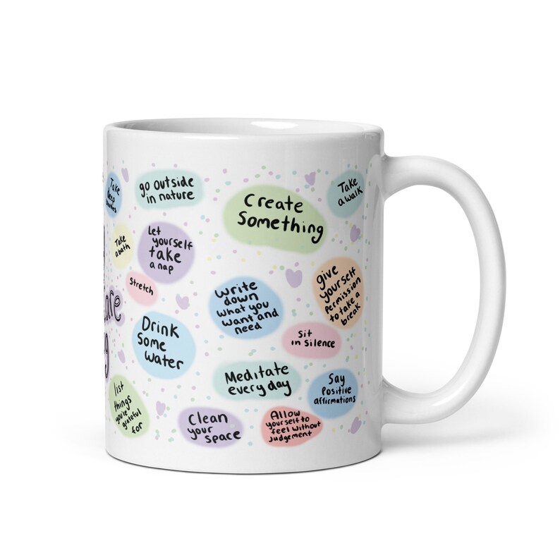 Peut inclure: Une tasse en c&eacute;ramique blanche avec un motif color&eacute; comportant des mots d'encouragement et des conseils d'auto-soins. Le texte comprend des phrases comme "Cr&eacute;er quelque chose", "Boire de l'eau", "M&eacute;diter tous les jours" et "Donnez-vous la permission de faire une pause".
