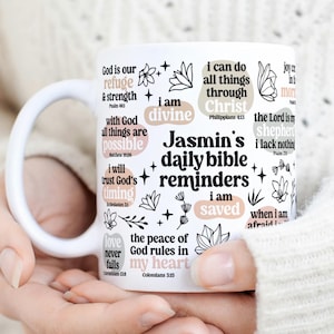 Könnte beinhalten: Weiße Keramiktasse mit schwarzem und rosafarbenem Text und floralen Designs. Der Text enthält Bibelverse und den Satz "Jasmin's daily bible reminders".