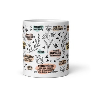 May include: White ceramic mug with black line art of flowers, leaves, and birds. The mug features inspirational phrases like "move your body," "rest," and "repeat positive affirmations" in various colors. A motivational gift.