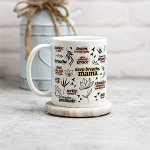 May include: White ceramic mug with a curved handle, featuring black text and floral illustrations. The mug includes phrases like "deep breaths, mama" and "practice gratitude." It rests on a round, light-colored coaster.