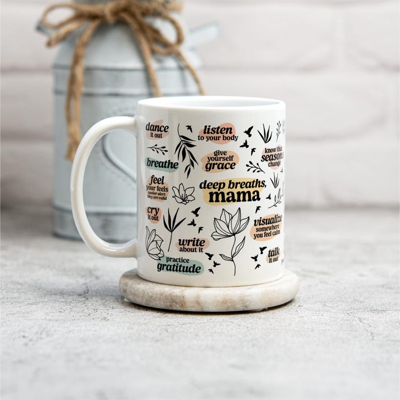 May include: White ceramic mug with a curved handle, featuring black text and floral illustrations. The mug includes phrases like "deep breaths, mama" and "practice gratitude." It rests on a round, light-colored coaster.