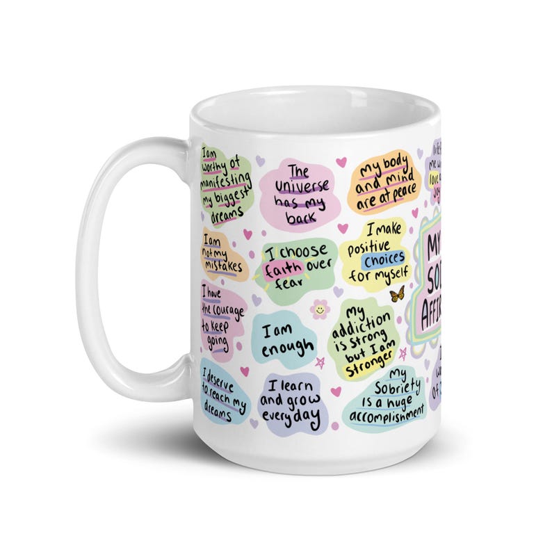 May include: White ceramic mug with colourful text and illustrations. The text includes positive affirmations such as "I am worthy of manifesting my biggest dreams", "The Universe has my back", "I choose faith over fear", "I am enough", "I learn and grow everyday", "My body and mind are at peace", "I make positive choices for myself", "My addiction is strong but I am stronger", "My sobriety is a huge accomplishment".