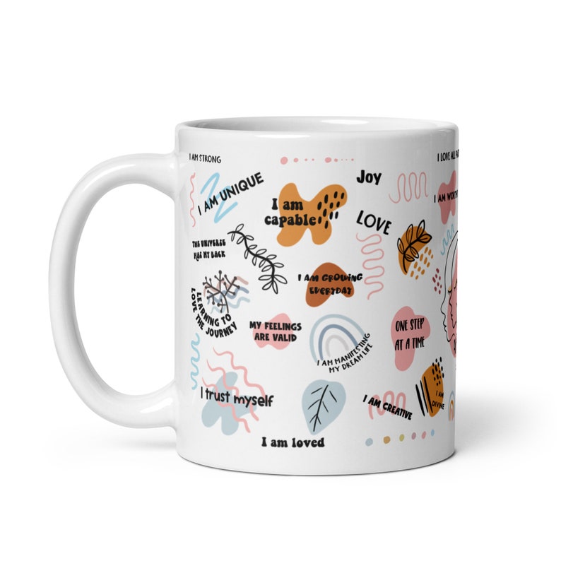 May include: White ceramic mug with a colorful design featuring positive affirmations and abstract shapes. The text includes phrases like "I am unique", "I am capable", "I am loved", and "I am growing everyday".
