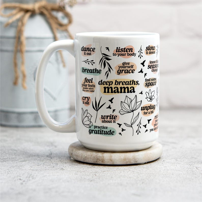 May include: White ceramic mug with a large handle, resting on a round stone coaster. The mug is decorated with black botanical illustrations and phrases like "deep breaths, mama" and "practice gratitude."