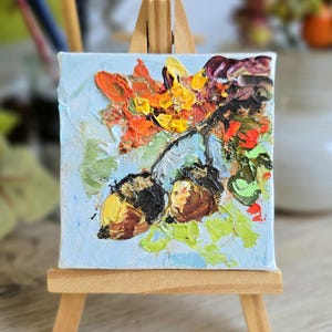 Puede incluir: Una pequeña pintura sobre lienzo cuadrado en un caballete de madera que presenta una pintura al óleo con técnica de impasto de dos bellotas marrones colgando de una rama con hojas de otoño naranjas, amarillas y verdes sobre un fondo azul claro.