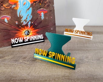 May include: Three different colored vinyl record stands with the words "NOW SPINNING" in gold lettering. The stands are white, green, and black.
