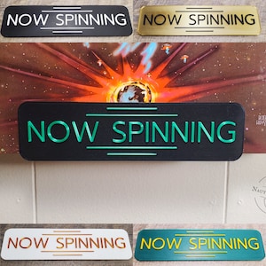 May include: Four different versions of a black sign with white, gold, green, and copper lettering that reads "NOW SPINNING".