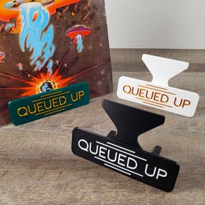 May include: Three vinyl record holders in green, black, and white with the words "QUEUED UP" in gold, white, and gold lettering, respectively. The holders are on a wood surface.