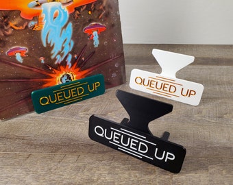 May include: Three vinyl record holders in green, black, and white with the words "QUEUED UP" in gold, white, and gold lettering, respectively. The holders are on a wood surface.