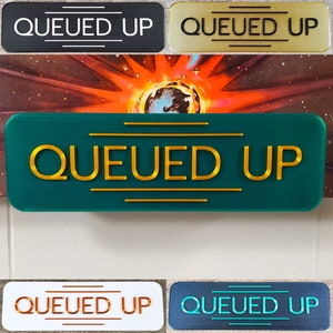 May include: Five different versions of a sign that reads "QUEUED UP" in gold letters. The signs are made of different materials and colors, including black, gold, green, white, and blue.