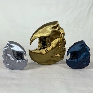 May include: Three 3D printed sculptures in the shape of a skull. The largest sculpture is gold, the middle sculpture is silver, and the smallest sculpture is blue.