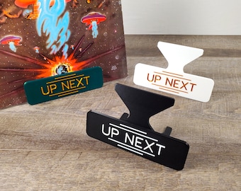 May include: Three different colored 'UP NEXT' signs. The signs are made of metal and have a modern design. The signs are perfect for displaying on a record player or turntable.