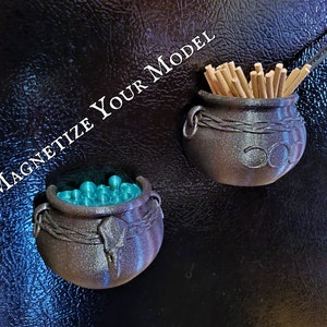A pair of witch cauldron magnets on a refrigerator door with the nearby text "Magnetize Your Model" indicate that commissioned custom 3D printing projects can include hardware such as magnets.
