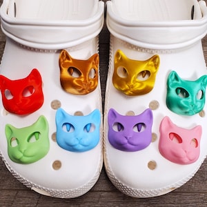 May include: A pair of white clogs with eight colorful cat-shaped shoe charms. The charms are in shades of red, orange, yellow, green, blue, purple, and pink.