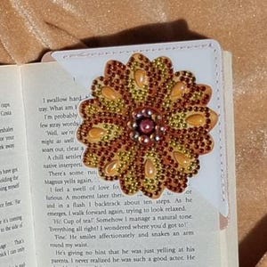 Op de afbeelding: Een close-up van een boek met een kleurrijk kralenbloem-bladwijzer. De bladwijzer is gemaakt van gele, oranje en rode kralen en heeft een bruin midden. Het boek is open op een pagina met tekst over een bruiloft.