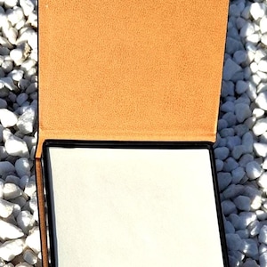 Può includere: Un porta bloc notes in pelle nera con una copertina in pelle marrone e un bloc notes bianco. Il porta bloc notes è appoggiato su un letto di ciottoli bianchi.