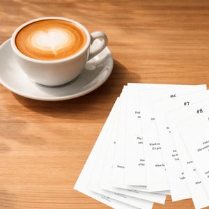 Könnte beinhalten: Eine weiße Kaffeetasse mit Latte-Art in Herzform auf einer weißen Untertasse. Ein Stapel weißer Karten mit Text und Zahlen liegt auf einer Holzoberfläche.
