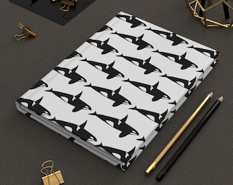 May include: A hardcover journal with a repeating pattern of black and white orca whales on a light gray background. Gold and black pencils, gold binder clips, and a geometric gold object are also present.