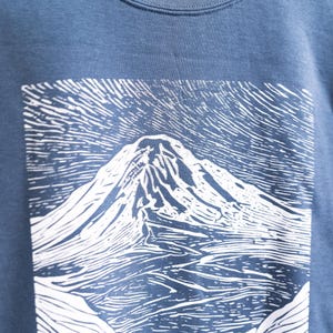 May include: Blue sweatshirt featuring a white graphic of a mountain landscape. The design is a detailed, linocut-style print. The sweatshirt is made of a soft, textured fabric.