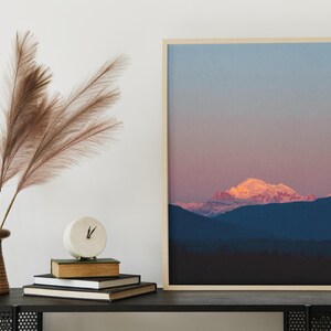 Puede incluir: Una impresión enmarcada de una cordillera al atardecer. Las cumbres de las montañas están cubiertas de nieve y el cielo es de un suave rosa y azul. La impresión está colgada en una pared blanca sobre una consola negra con un jarrón de hierba de la pampa seca y un reloj.