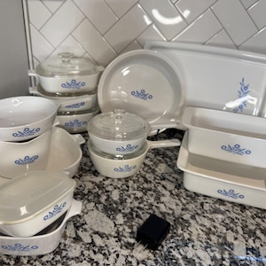 May include: A collection of white ceramic bakeware and cookware pieces featuring a blue floral pattern. The set includes various rectangular, square, and round dishes, some with clear glass lids, a round skillet, and a large rectangular serving tray.
