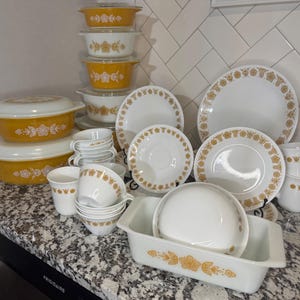 May include: A collection of white and yellow glass dinnerware featuring a repeating floral pattern. The set includes lidded casserole dishes, various sizes of bowls, plates, saucers, and mugs, all decorated with a consistent gold-toned flower motif.