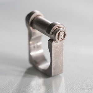 Può includere: Un anello in metallo argentato con un design quadrato e una piccola lettera "A" incisa sulla parte superiore.