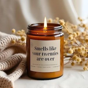 Könnte beinhalten: Eine brennende Kerze in einem braunen Glas. Auf dem Etikett steht "Smells like your twenties are over". Die Kerze ist von getrockneten Blumen und einer beigen Strickdecke umgeben. Die Kerze wiegt 180g.
