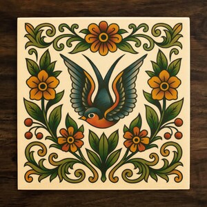 May include: Square ceramic tile featuring a traditional tattoo-style design. A blue and orange swallow with outstretched wings is centered, surrounded by orange flowers, green leaves, and decorative flourishes. The tile has a cream-colored background.