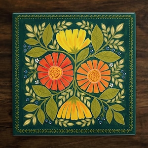May include: Square ceramic tile featuring a floral design. The artwork showcases orange and yellow flowers with green leaves and blue berries against a dark green background. The tile is framed with a dark green border and is set on a wooden surface.