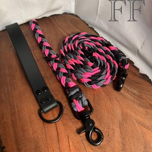 May include: A black leather dog collar and a matching leash. The leash is braided with pink, gray, and black paracord. The collar has a black metal D-ring and the leash has a black metal clasp. The items are displayed on a wooden surface.