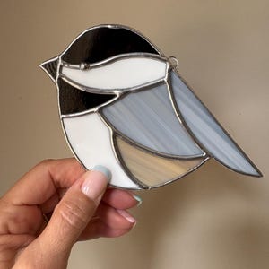 May include: Stained glass bird with a black head, white chest, and gray wings. The bird has a silver outline and a small loop for hanging. The bird's body has a tan and white section. A decorative piece for home decor.